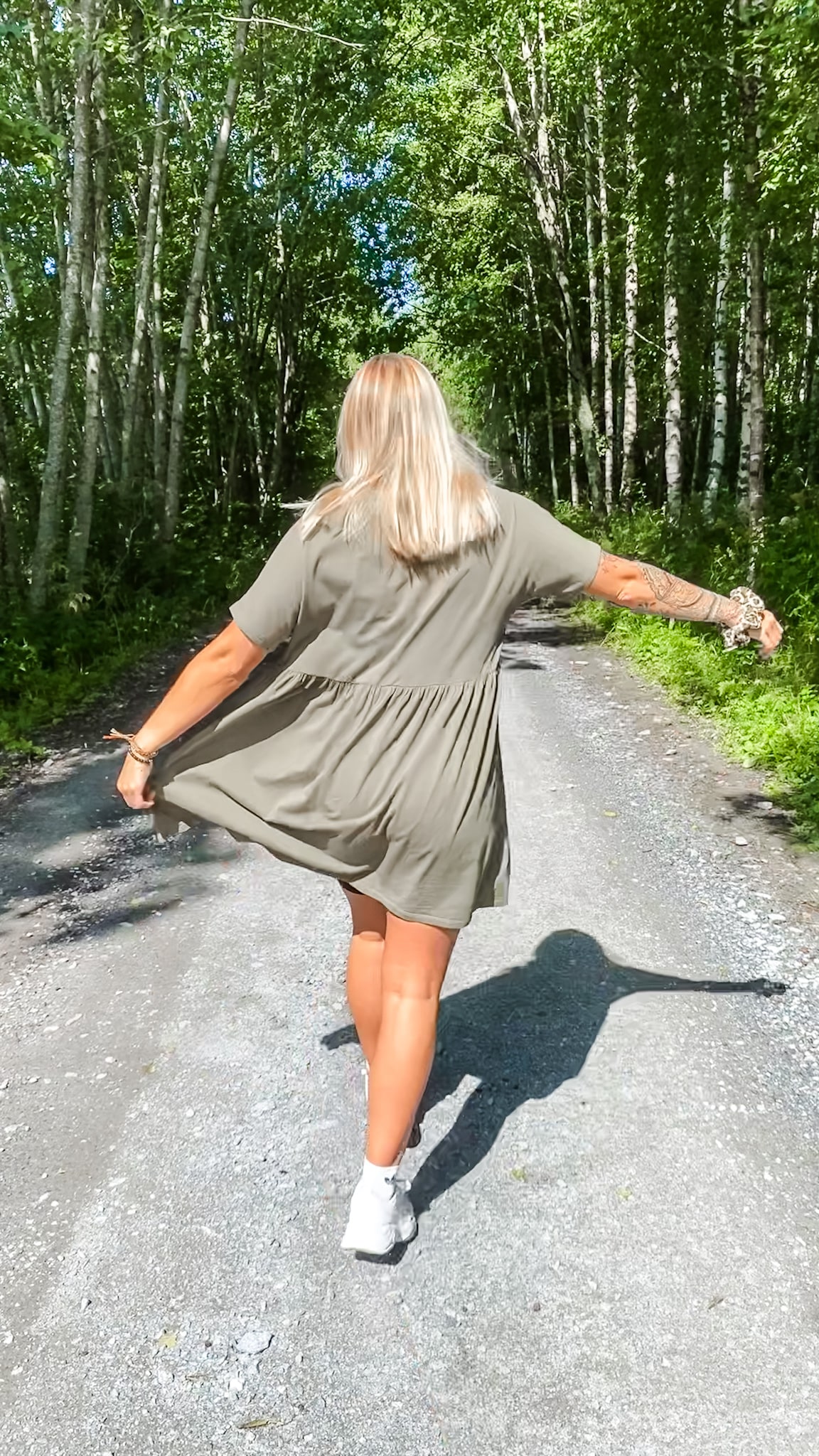 Kesäisessä maisemassa vaaleatukkainen nainen mekko päällä kävelemässä tiellä selkä kameraan päin